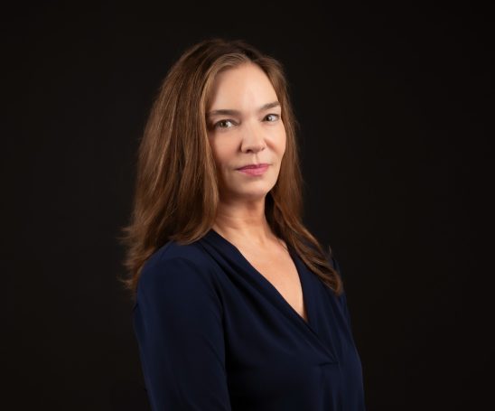 Headshot of a professional executive woman with long brown hair & a blue blouse poses against a dark background