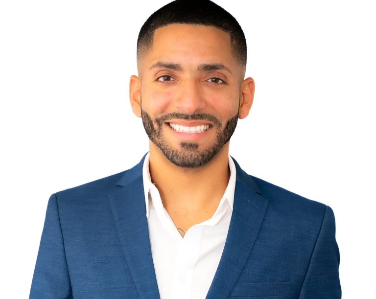 Man in a suit and sunglasses, smiling, outdoors with blurred background photographed by Iris Ramil Photographer.