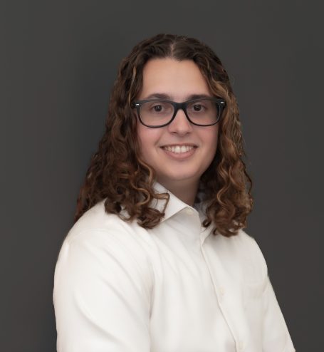 Headshot of young professional man with long curly hair, wearing glasses & a white shirt against a dark background.
