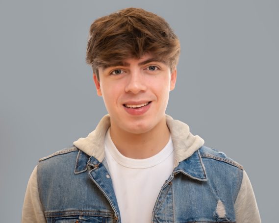 Young model at Headshot Studio Portrait studio with brown hair wearing a denim jacket and a white shirt, standing against a gray background.