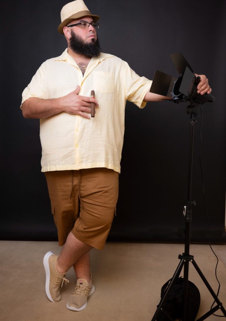 Man in a yellow shirt and hat poses next to a microphone stand against a dark background.