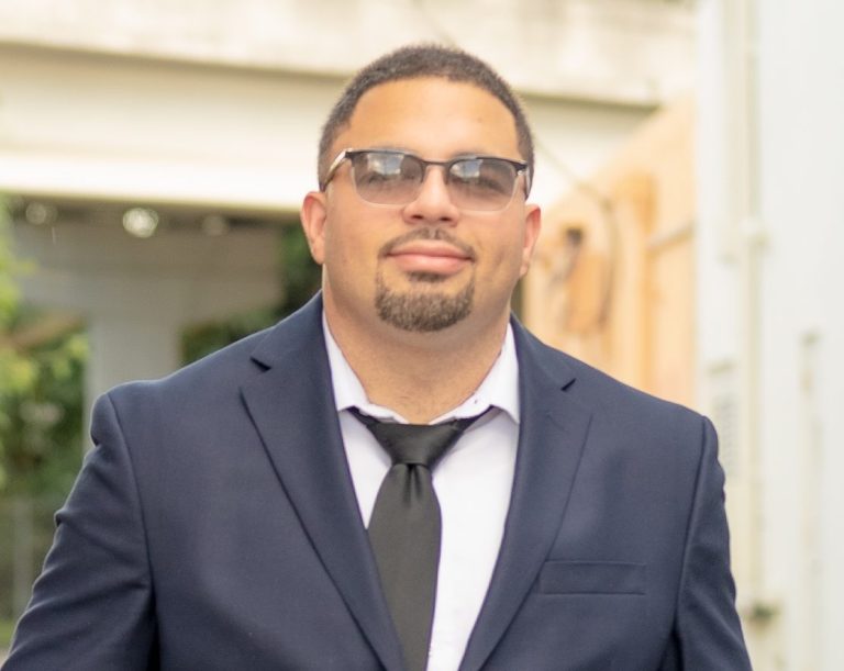 Man in a suit and sunglasses, smiling, outdoors with blurred background photographed by Iris Ramil Photographer.